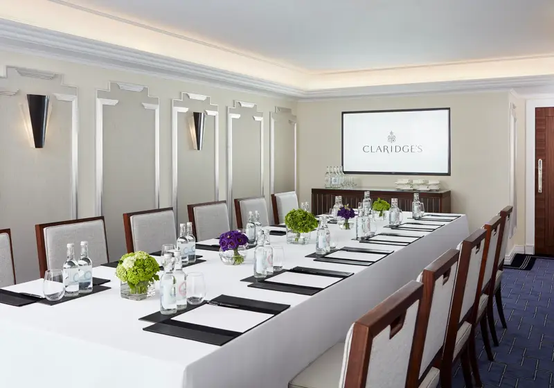 Modern meeting room at Claridge’s arranged with a long conference table, elegant chairs, bottled water, notepads, and a screen displaying the hotel’s logo.