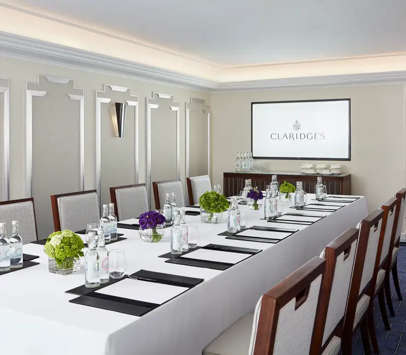 Modern meeting room at Claridge’s arranged with a long conference table, elegant chairs, bottled water, notepads, and a screen displaying the hotel’s logo.