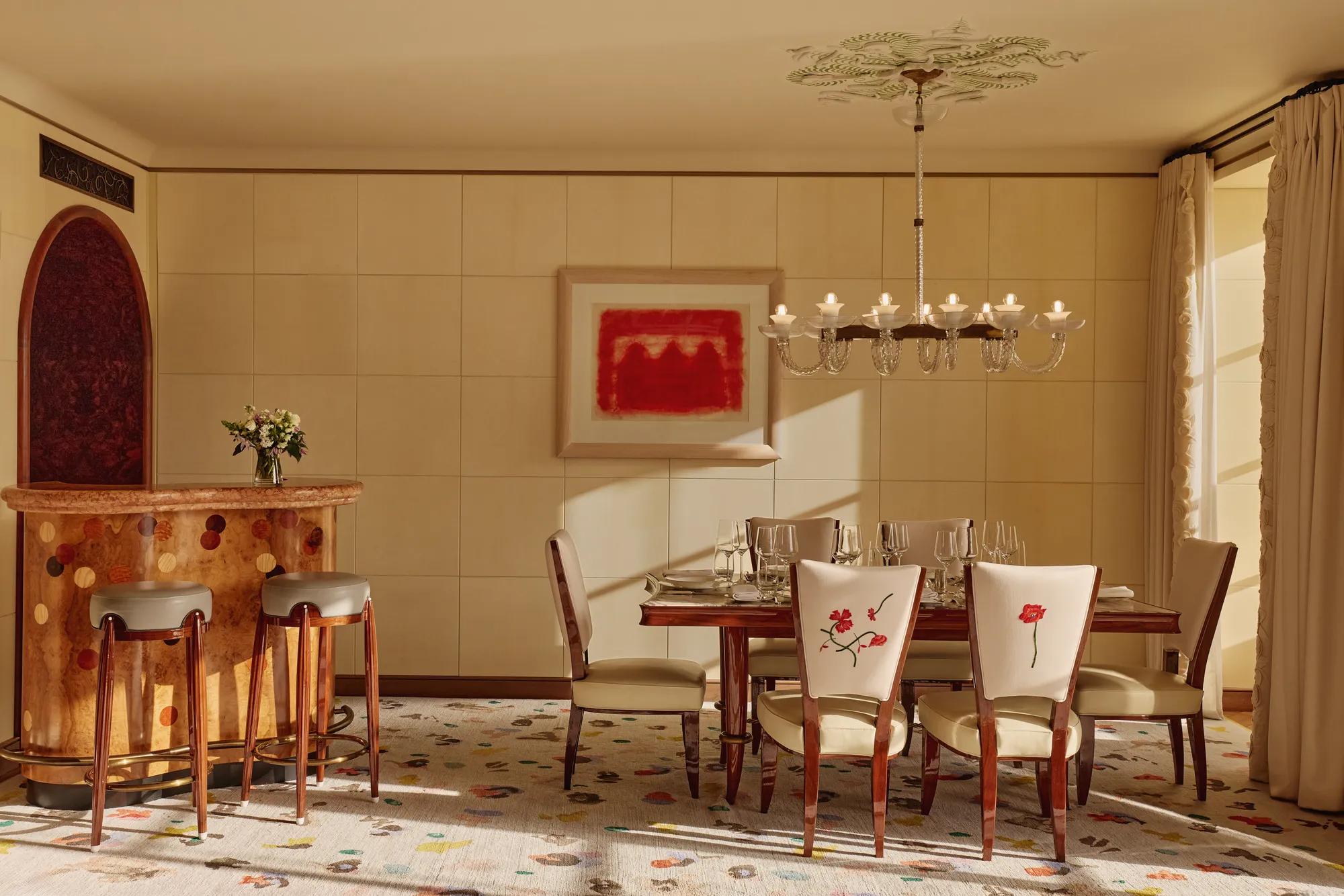 Elegant dining room with a wooden table set for six, cream chairs embroidered with red flowers, a chandelier overhead, and a curved wooden bar with stools, all softly lit by natural daylight.
