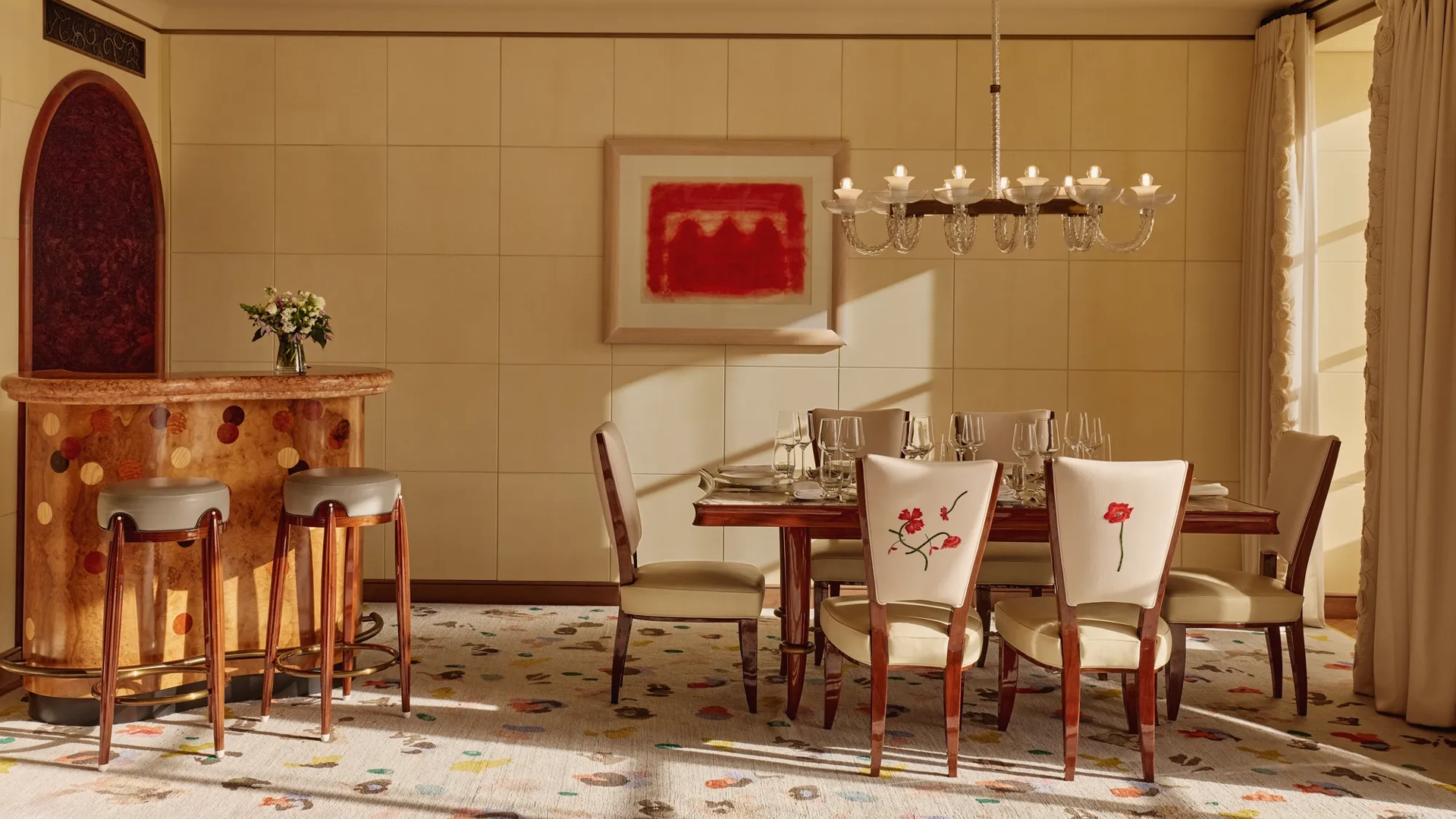 Elegant dining room with a wooden table set for six, cream chairs embroidered with red flowers, a chandelier overhead, and a curved wooden bar with stools, all softly lit by natural daylight.