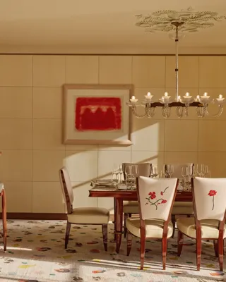 Elegant dining room with a wooden table set for six, cream chairs embroidered with red flowers, a chandelier overhead, and a curved wooden bar with stools, all softly lit by natural daylight.