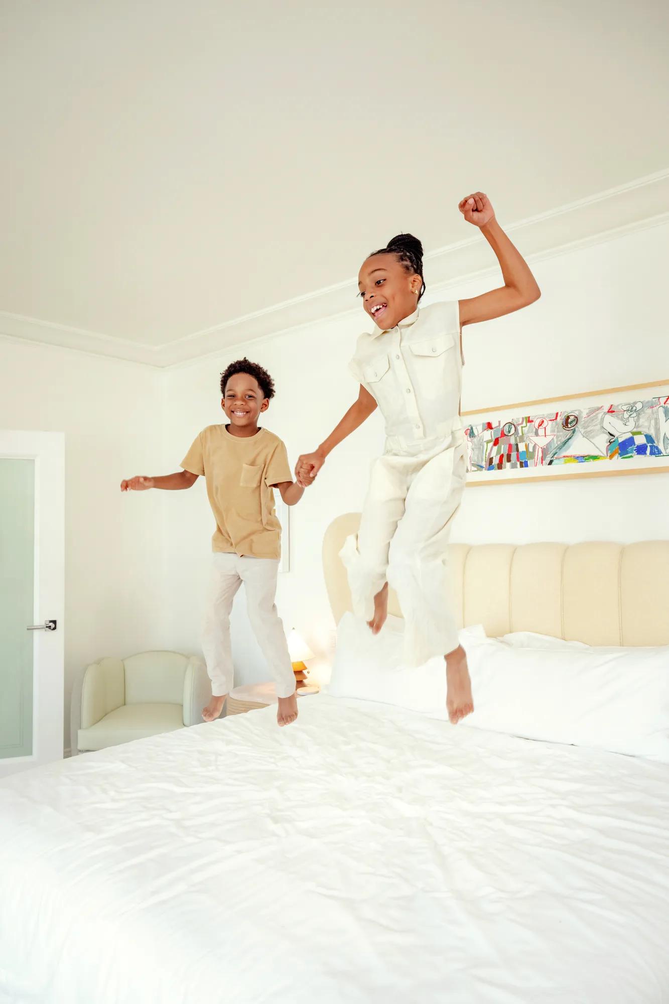 Two children holding hands, jumping on a hotel bed in a light-filled Hollywood suite