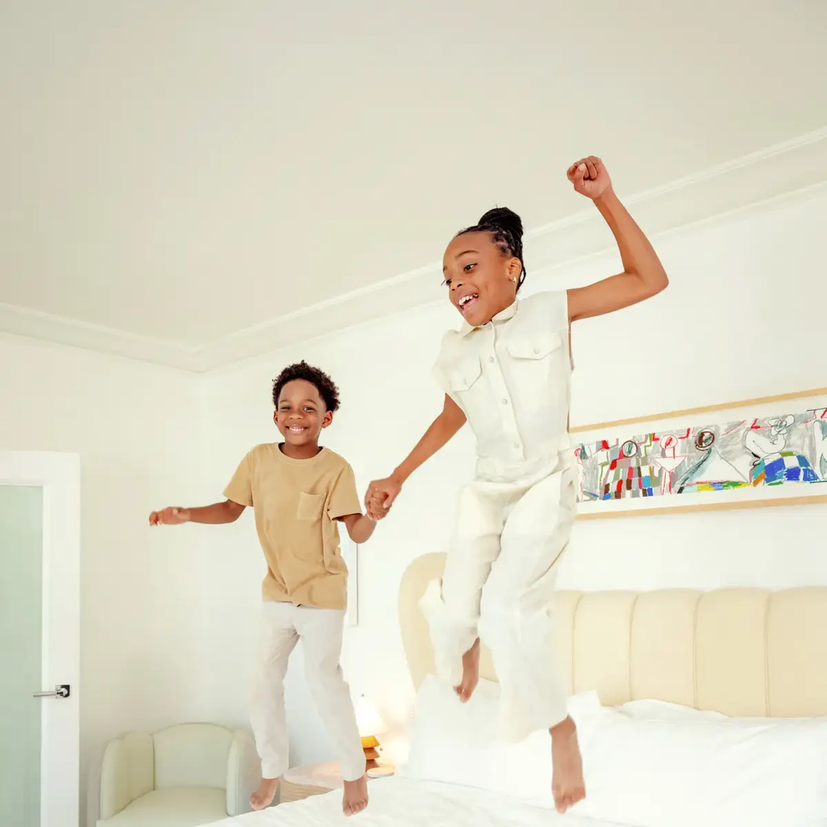 Two children holding hands, jumping on a hotel bed in a light-filled Hollywood suite