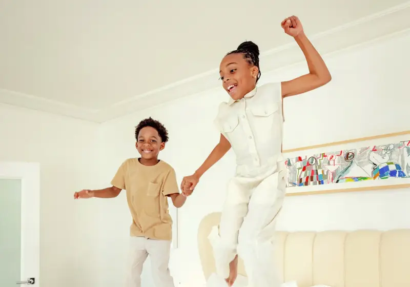 Two children holding hands, jumping on a hotel bed in a light-filled Hollywood suite