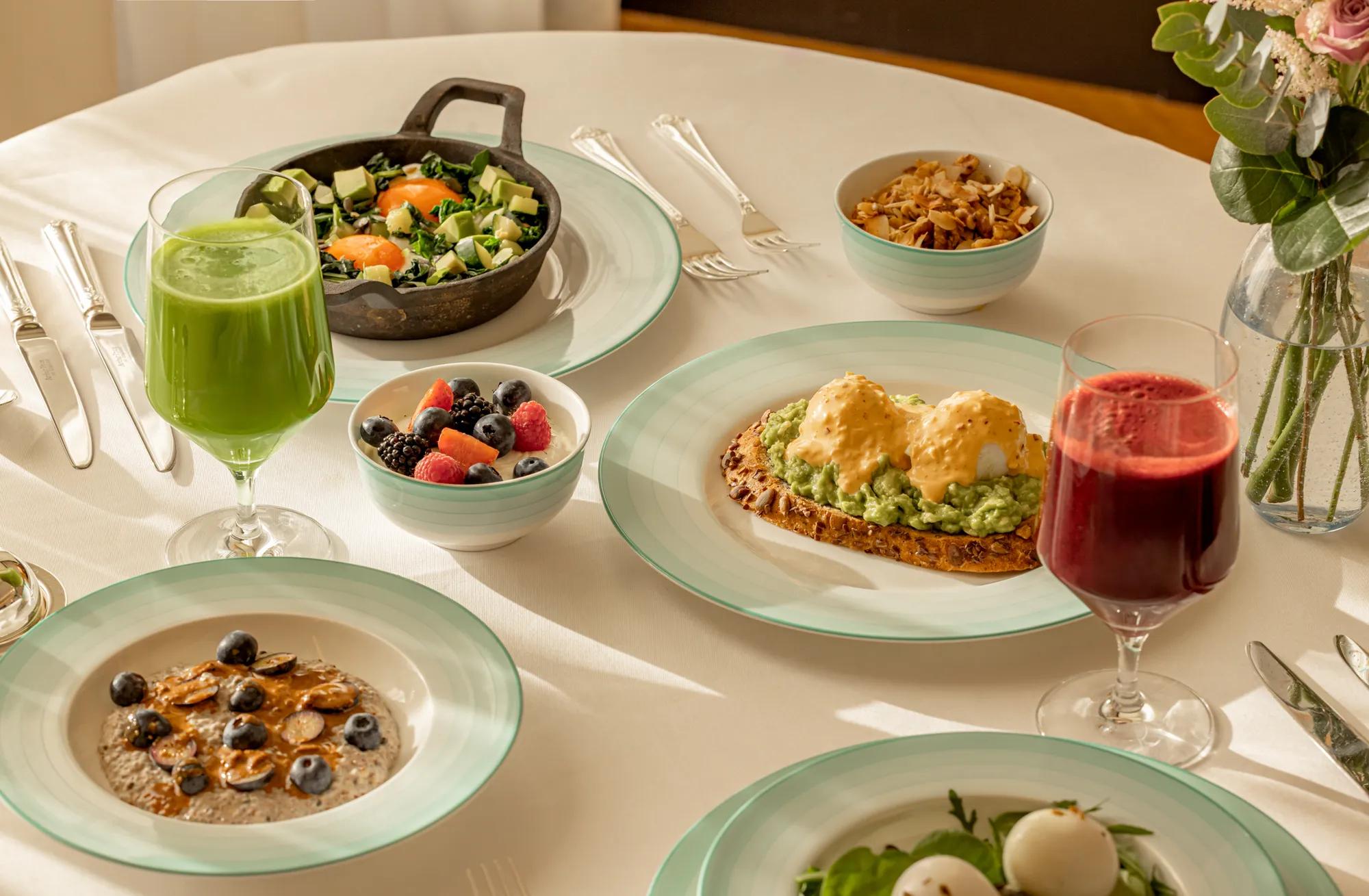 Breakfast table with eggs on toast, granola, fruit bowls, salads, porridge, and glasses of green and berry juice.