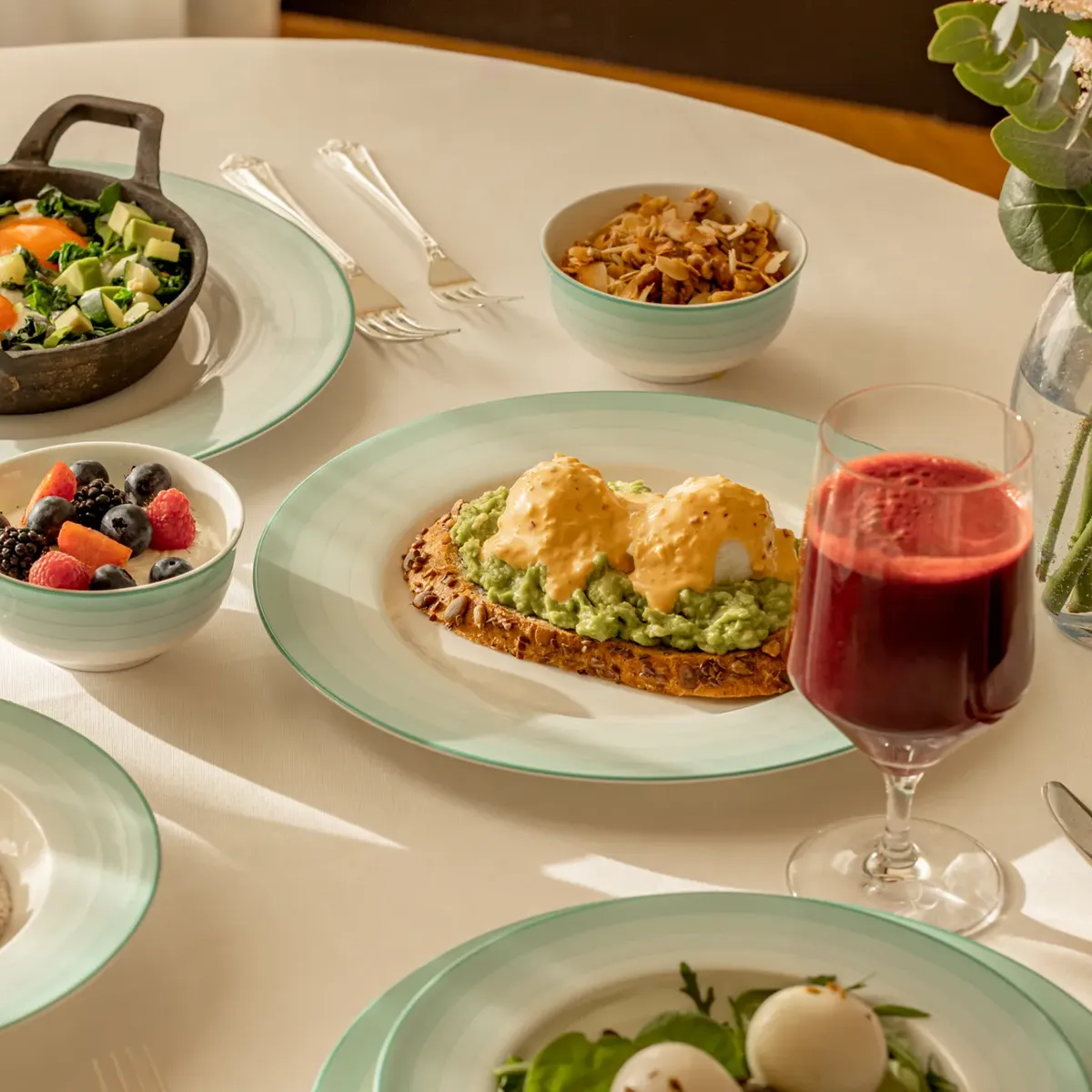 Breakfast table with eggs on toast, granola, fruit bowls, salads, porridge, and glasses of green and berry juice.