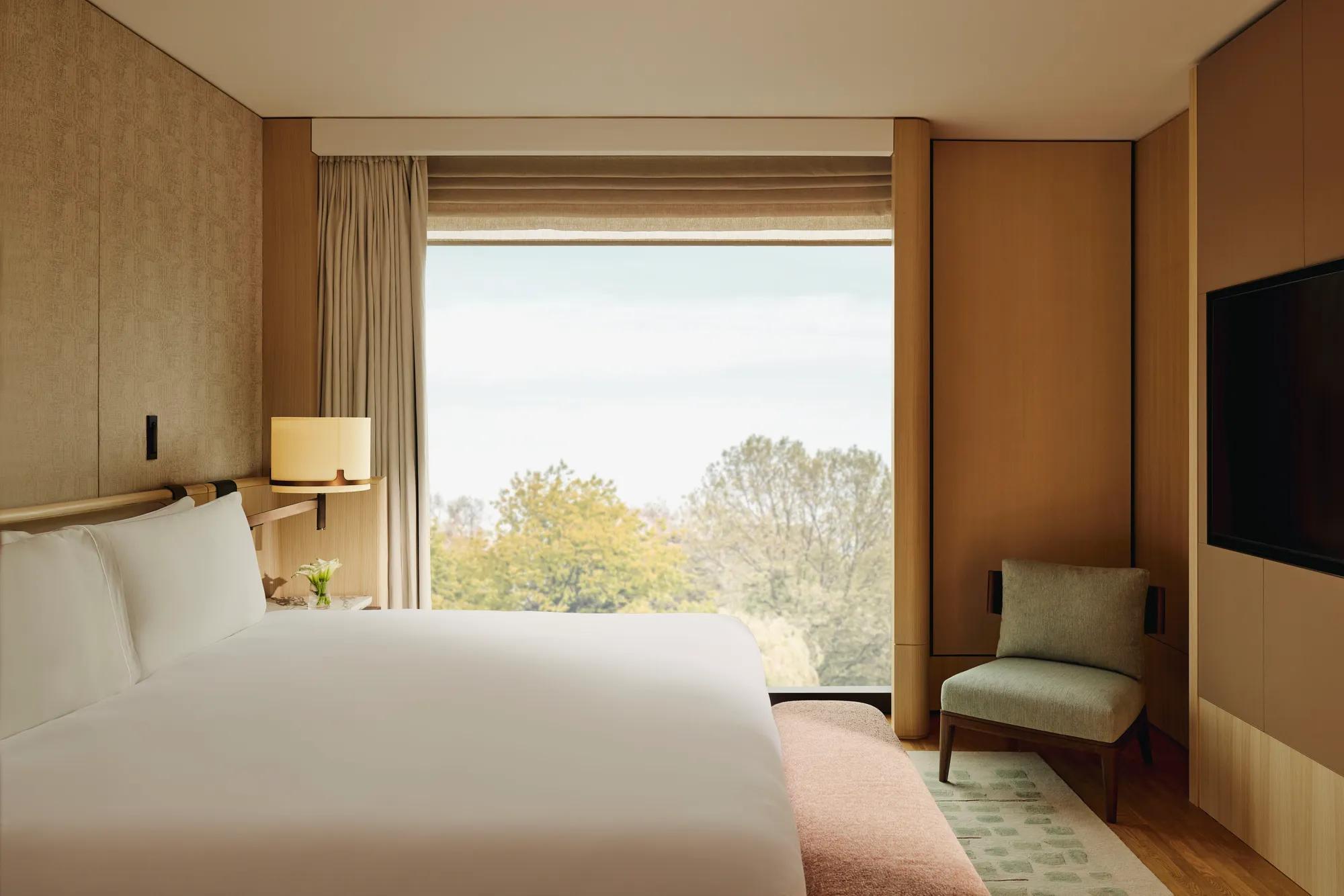 Modern hotel bedroom with king bed, soft wood panelling, armchair and floor-to-ceiling window overlooking leafy trees.