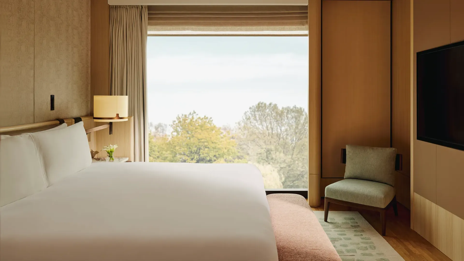 Modern hotel bedroom with king bed, soft wood panelling, armchair and floor-to-ceiling window overlooking leafy trees.