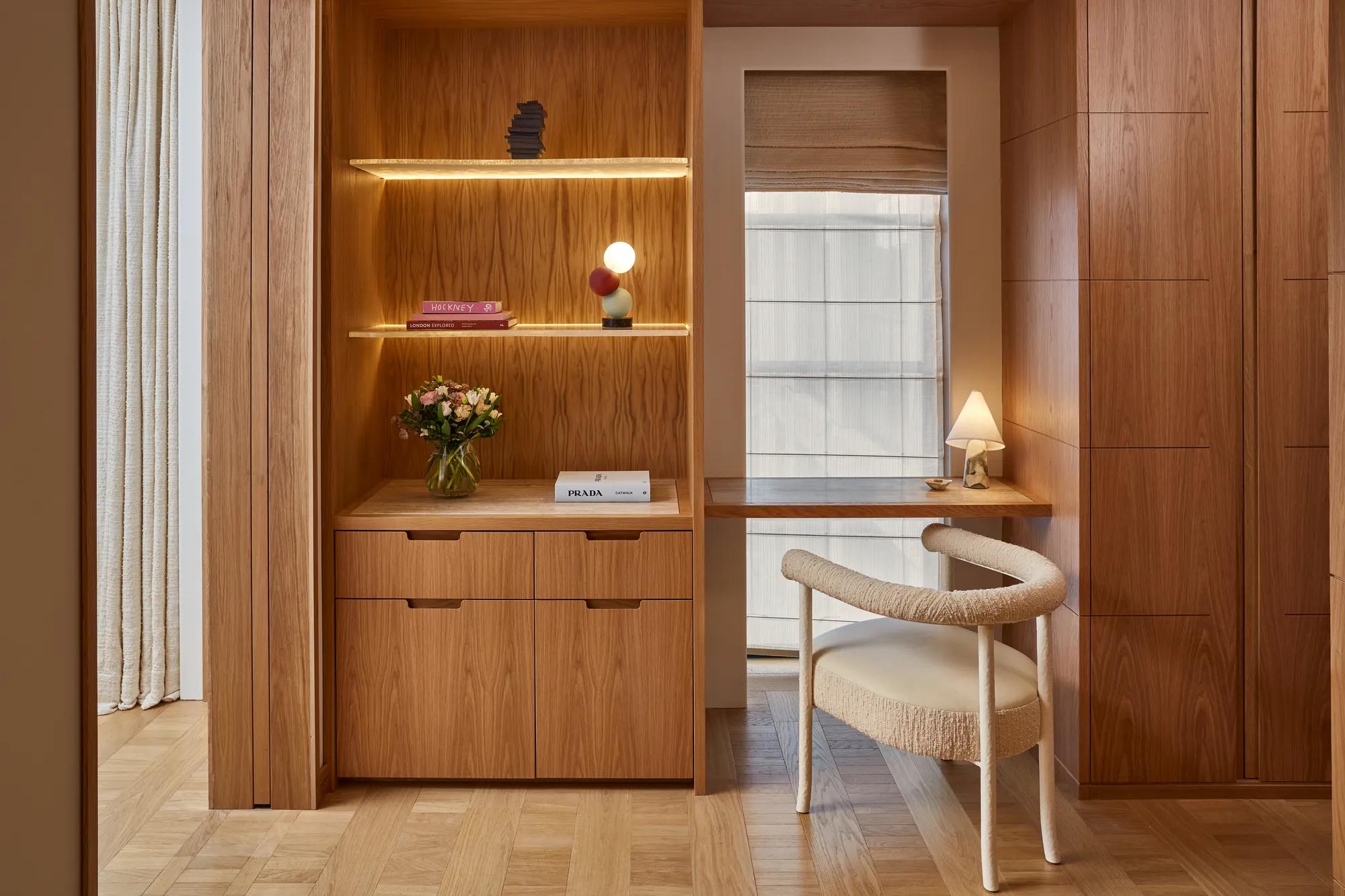 Wood-paneled workspace with built-in shelving and drawers, small desk by window, upholstered chair, and decorative lighting.
