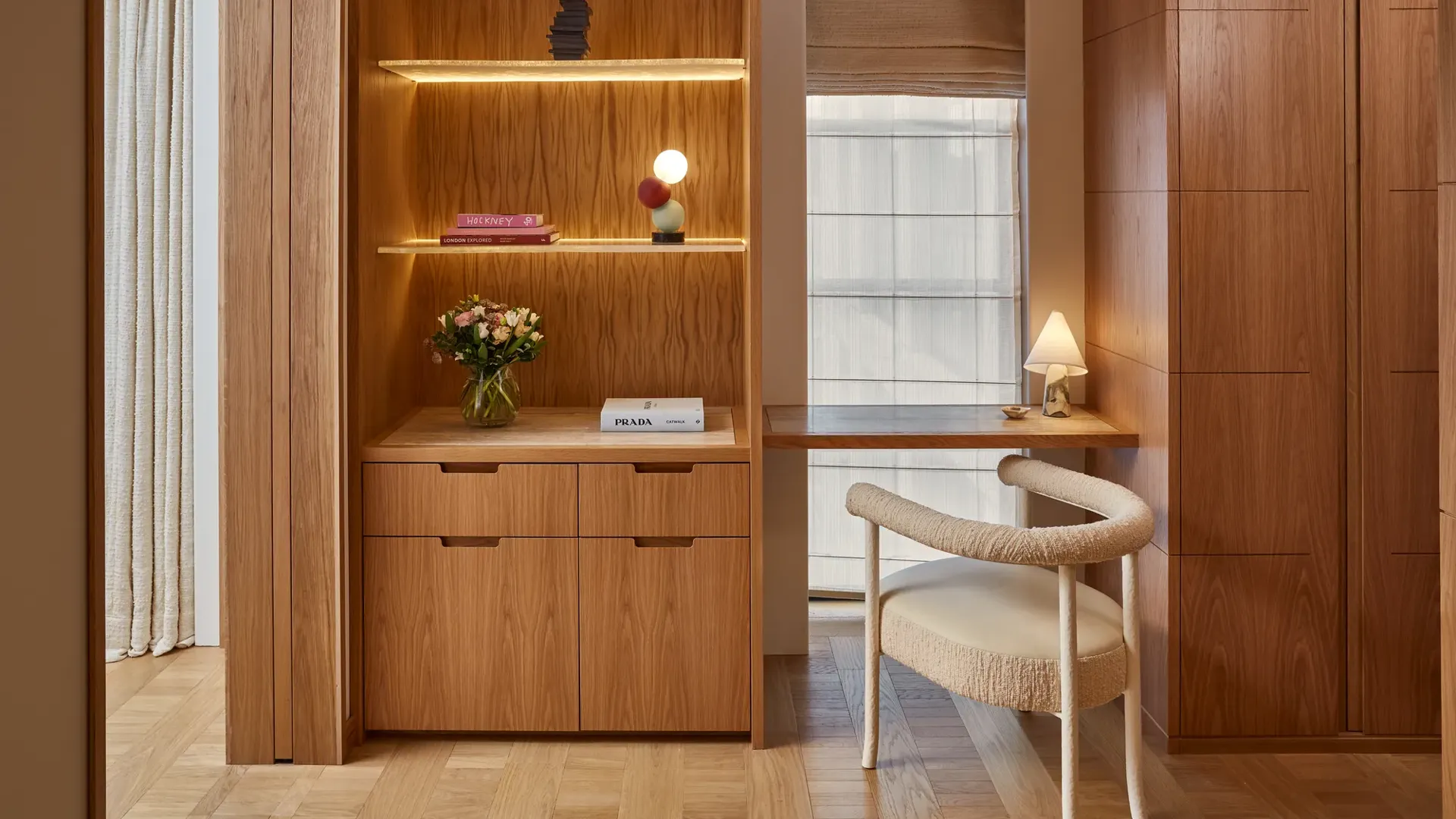 Wood-paneled workspace with built-in shelving and drawers, small desk by window, upholstered chair, and decorative lighting.