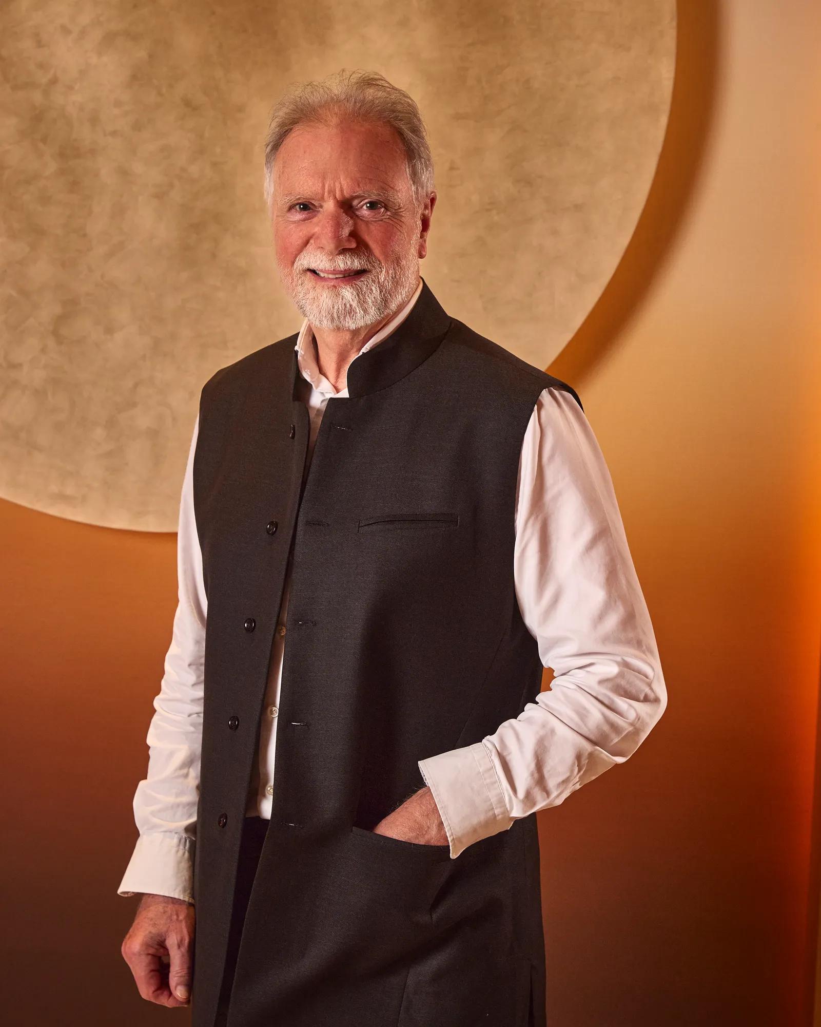 Smiling man with grey hair and beard in a dark vest and white shirt against a warm-toned background with a circular feature