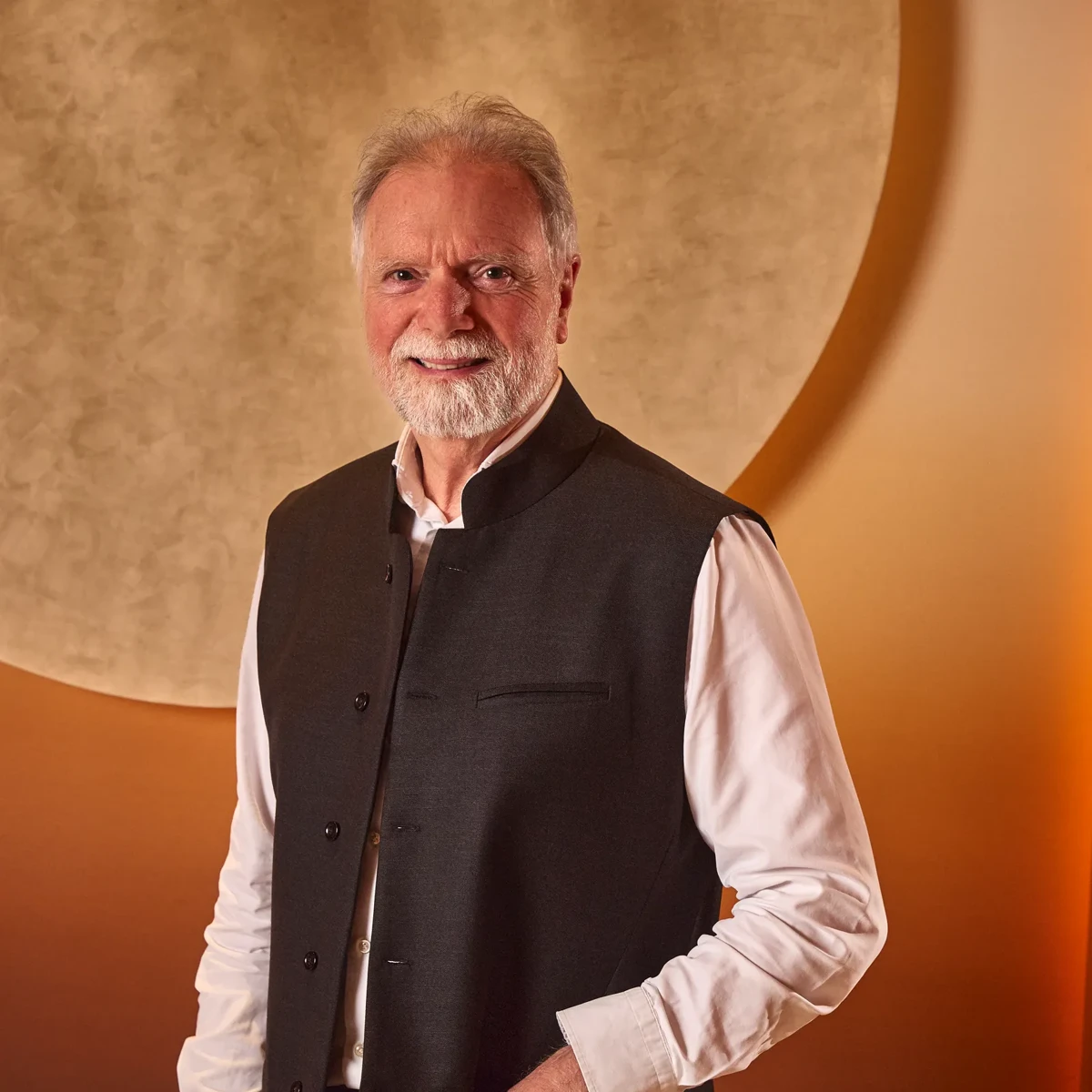 Smiling man with grey hair and beard in a dark vest and white shirt against a warm-toned background with a circular feature