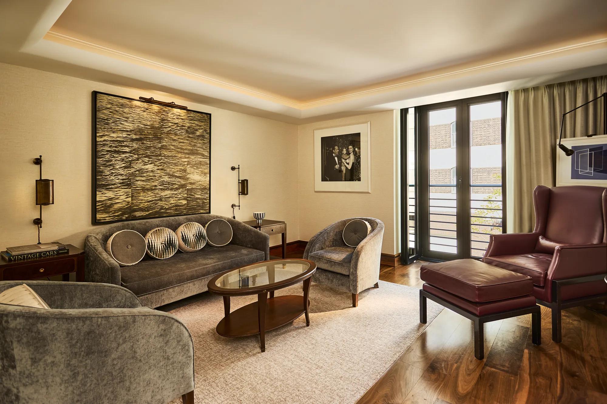 **Alt text:**
Elegant living room with grey velvet seating, walnut floors, curated artwork, a burgundy armchair and French doors opening to a private balcony.
