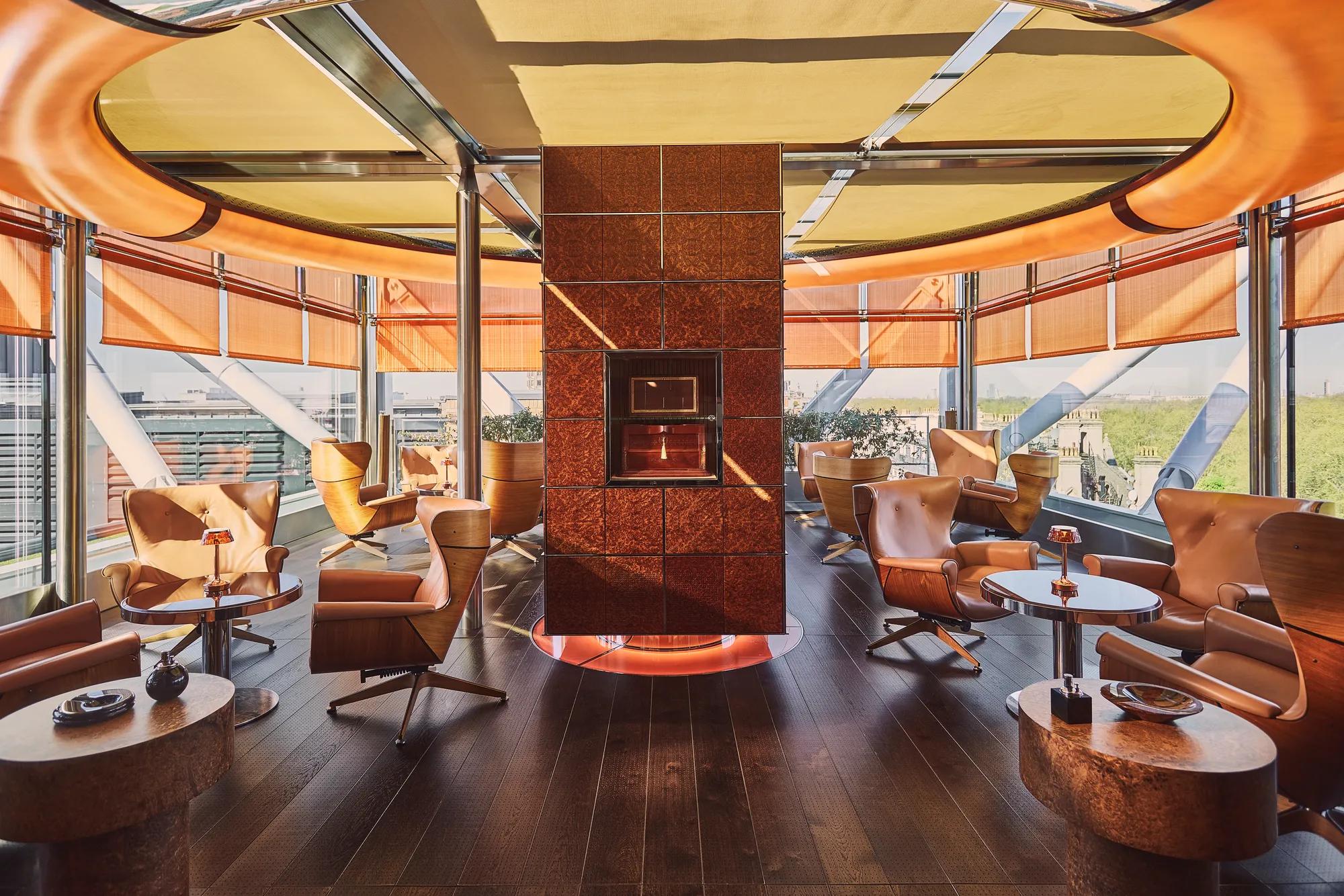 Circular rooftop lounge with leather armchairs, central fireplace, and panoramic city views through floor-to-ceiling windows.