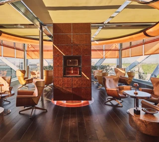 Circular rooftop lounge with leather armchairs, central fireplace, and panoramic city views through floor-to-ceiling windows.