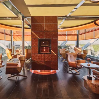 Circular rooftop lounge with leather armchairs, central fireplace, and panoramic city views through floor-to-ceiling windows.
