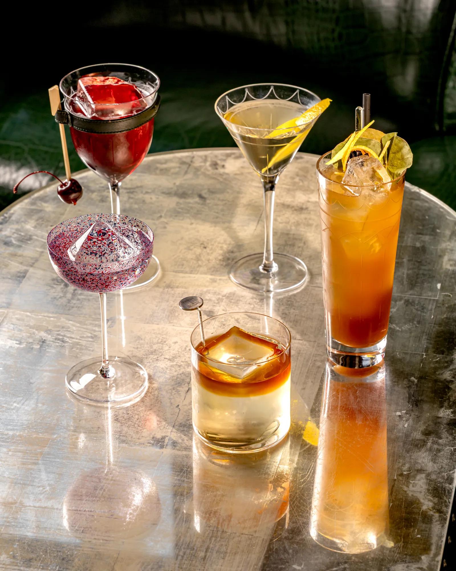 Assorted cocktails on reflective metal table, including martini, old fashioned, spritz, and red drink garnished with citrus and cherry.