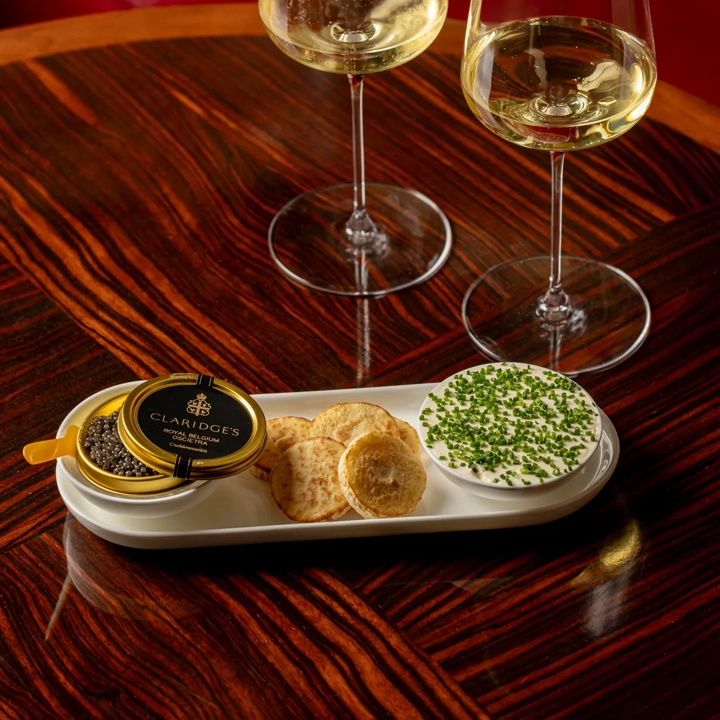 Two glasses of white wine with caviar, blinis, and chive-topped crème fraîche on a wooden table.