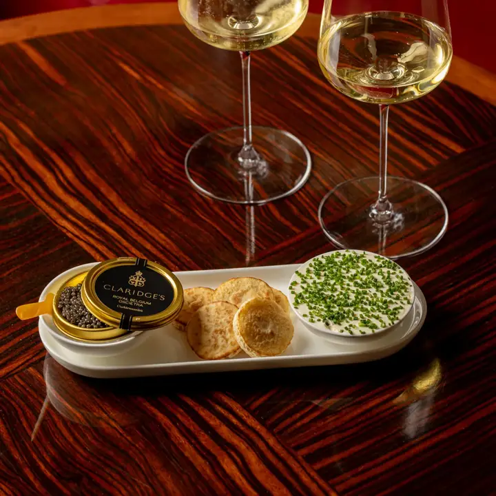 Two glasses of white wine with caviar, blinis, and chive-topped crème fraîche on a wooden table.