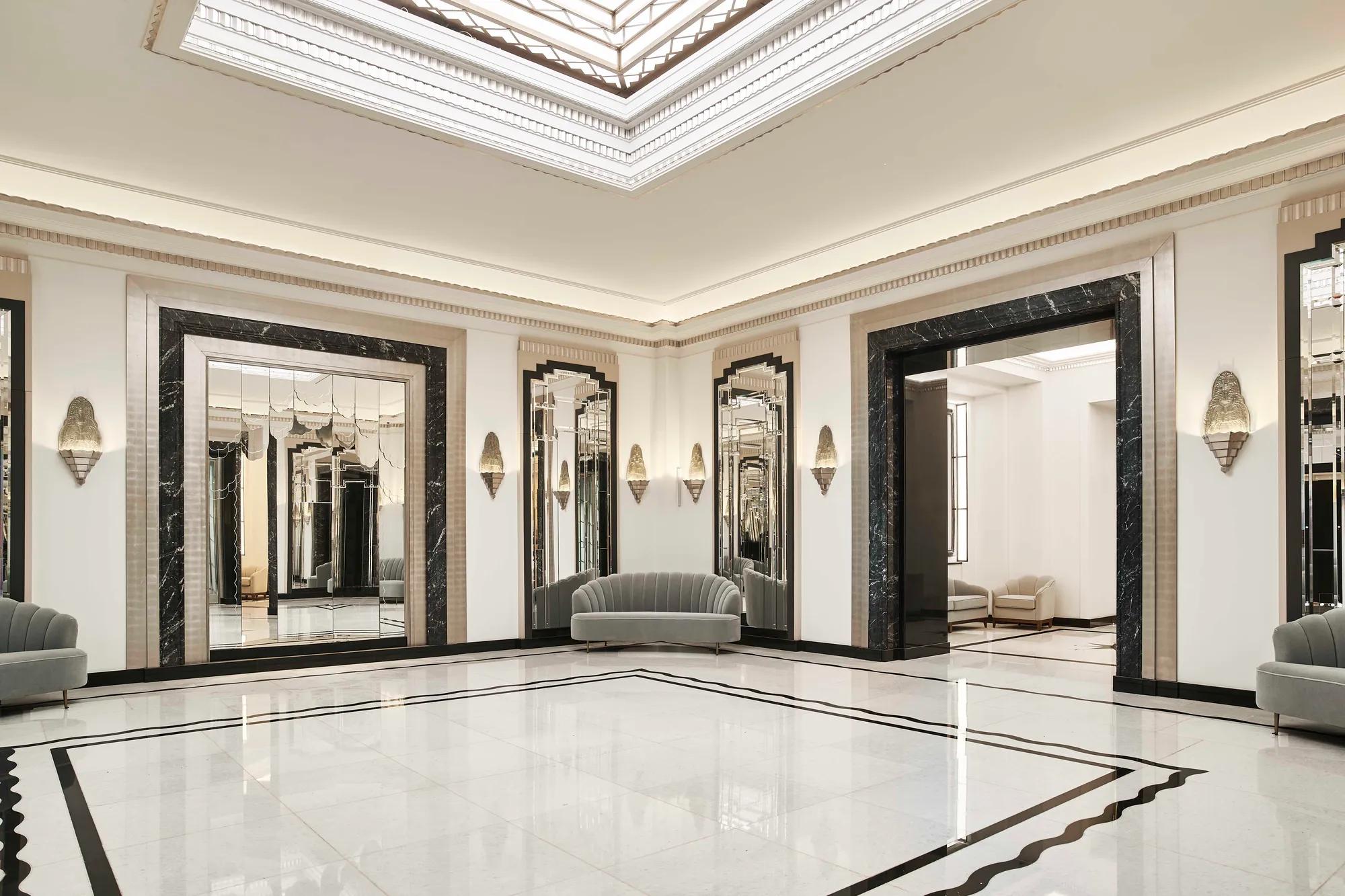 Spacious Art Deco foyer at Claridge’s featuring a geometric marble floor, mirrored walls framed in black marble, soft grey curved sofas, and an illuminated glass ceiling.
