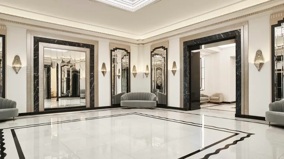 Spacious Art Deco foyer at Claridge’s featuring a geometric marble floor, mirrored walls framed in black marble, soft grey curved sofas, and an illuminated glass ceiling.