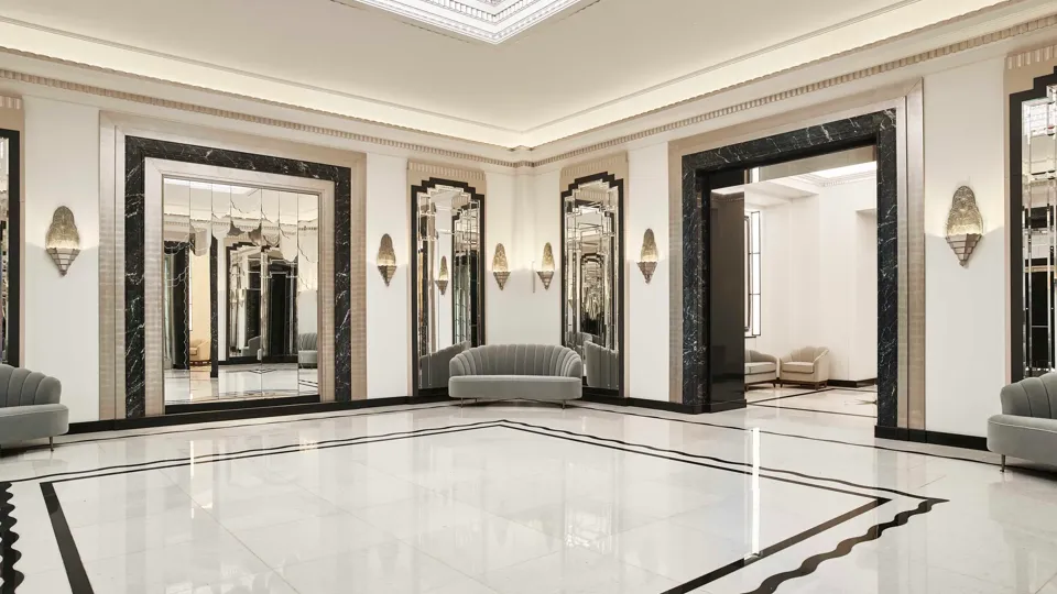 Spacious Art Deco foyer at Claridge’s featuring a geometric marble floor, mirrored walls framed in black marble, soft grey curved sofas, and an illuminated glass ceiling.