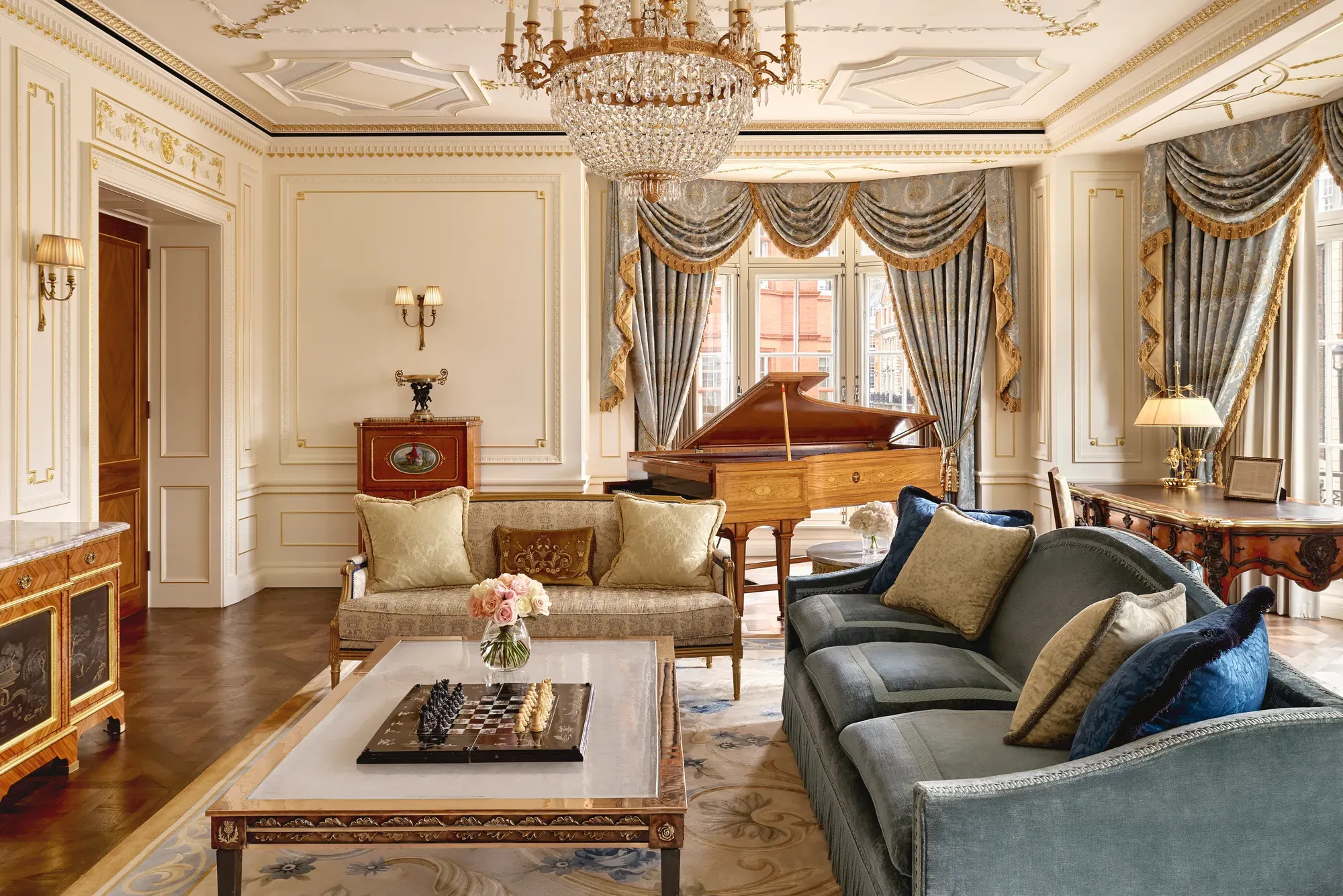 Grand living room with ornate gold detailing, blue and gold draped curtains, a crystal chandelier, plush sofas, and a polished wooden grand piano beside large windows.