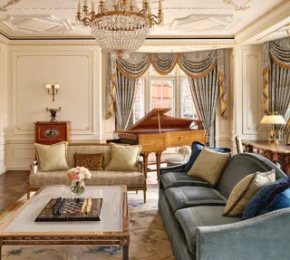 Grand living room with ornate gold detailing, blue and gold draped curtains, a crystal chandelier, plush sofas, and a polished wooden grand piano beside large windows.