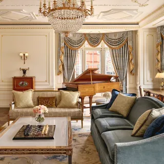 Grand living room with ornate gold detailing, blue and gold draped curtains, a crystal chandelier, plush sofas, and a polished wooden grand piano beside large windows.