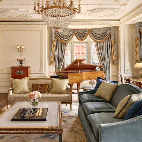 Grand living room with ornate gold detailing, blue and gold draped curtains, a crystal chandelier, plush sofas, and a polished wooden grand piano beside large windows.