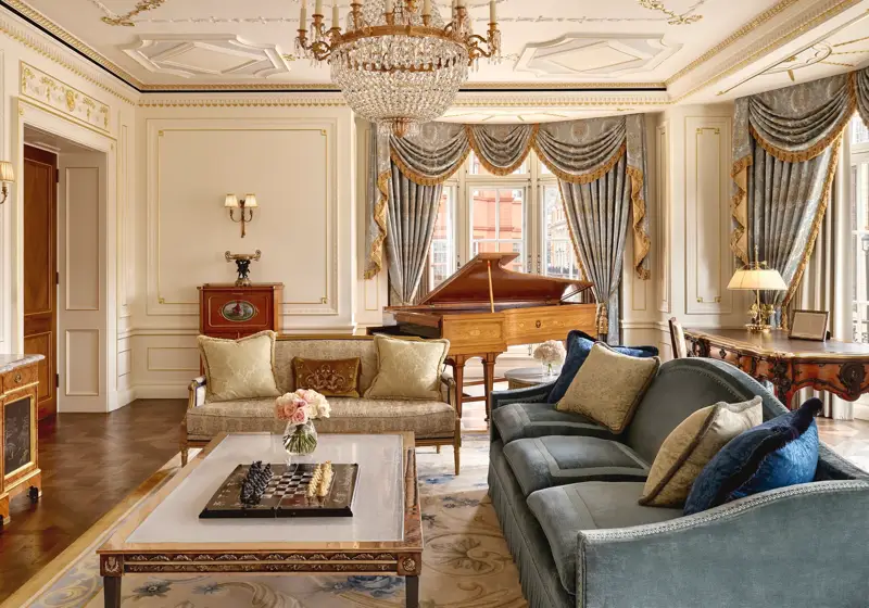 Grand living room with ornate gold detailing, blue and gold draped curtains, a crystal chandelier, plush sofas, and a polished wooden grand piano beside large windows.