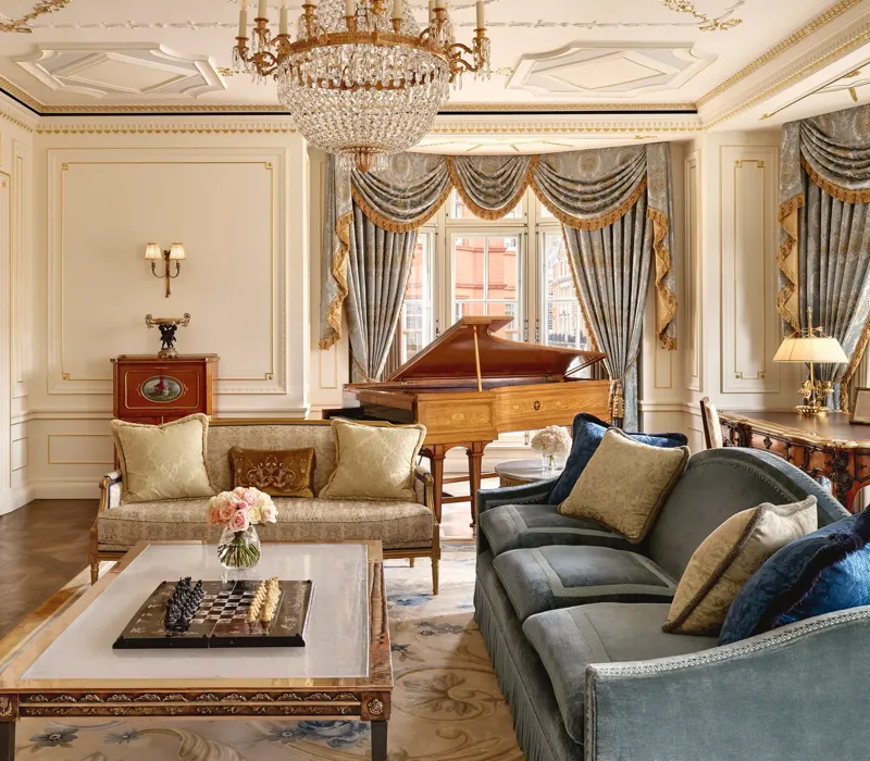 Grand living room with ornate gold detailing, blue and gold draped curtains, a crystal chandelier, plush sofas, and a polished wooden grand piano beside large windows.