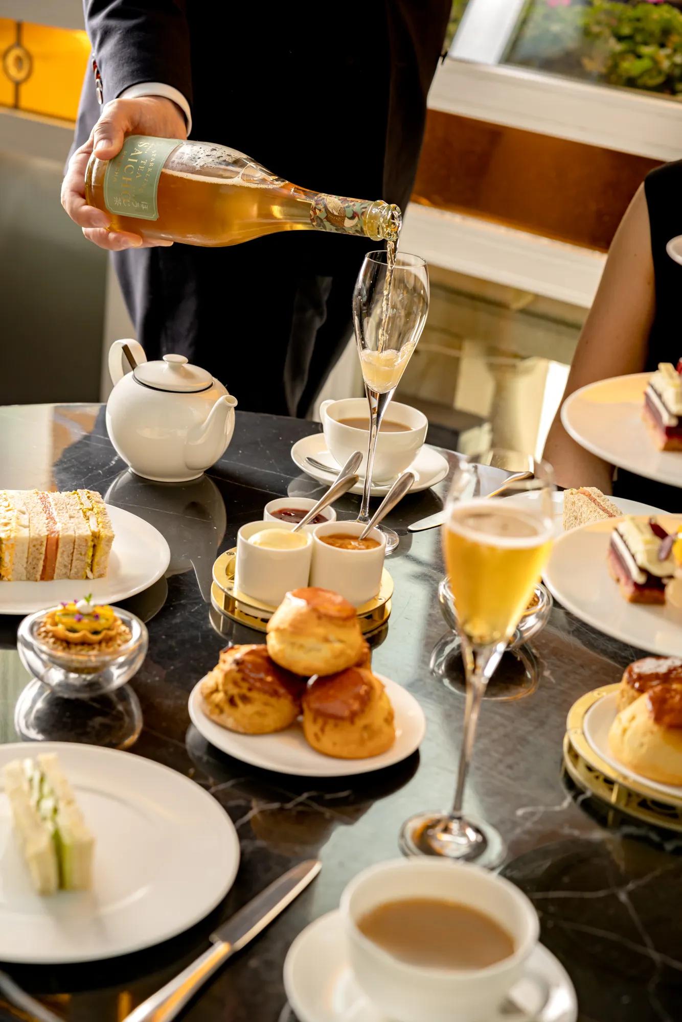 **Alt text:**
Champagne is poured into a flute beside a traditional afternoon tea with finger sandwiches, scones, pastries and tea service.
