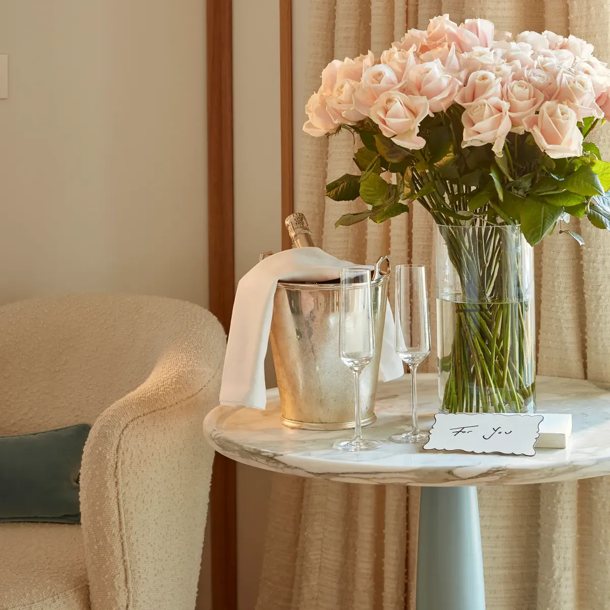 Vase of pale pink roses on a marble table with champagne, glasses, and a handwritten note in a softly lit suite.