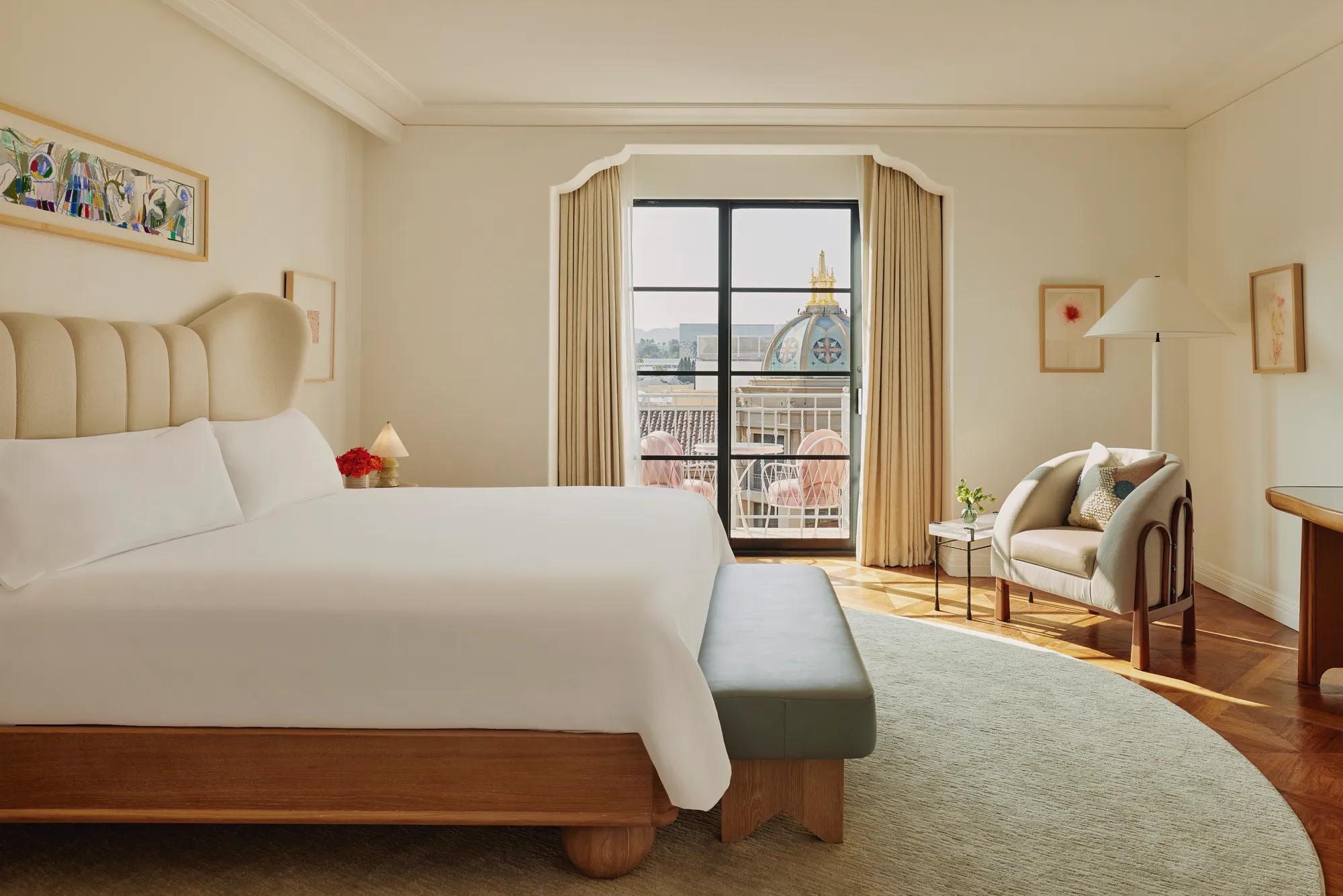 Elegant bedroom with a cream upholstered bed, grey bench, armchair with cushions, and balcony doors framing a view of a domed rooftop and pink terrace chairs.