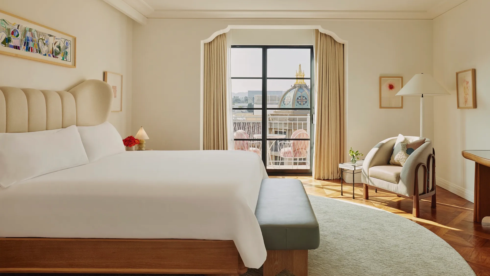 Elegant bedroom with a cream upholstered bed, grey bench, armchair with cushions, and balcony doors framing a view of a domed rooftop and pink terrace chairs.