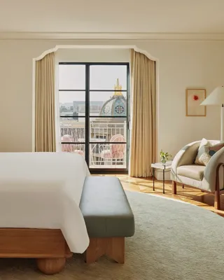 Elegant bedroom with a cream upholstered bed, grey bench, armchair with cushions, and balcony doors framing a view of a domed rooftop and pink terrace chairs.