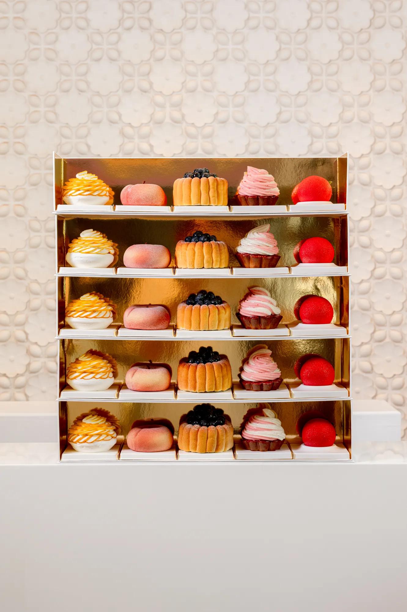 Gold display shelf presenting rows of miniature desserts, including meringues, peach-shaped pastries, blueberry cakes and strawberry-shaped sweets against a textured white backdrop.