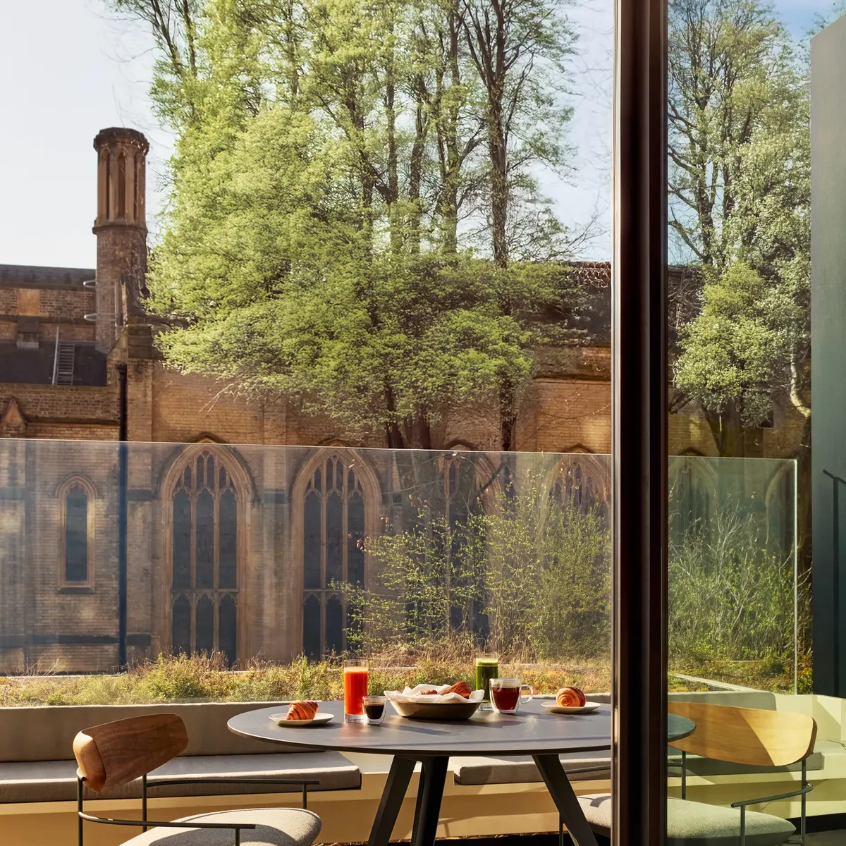 Outdoor table with croissants, juice, and coffee overlooking historic stone church and greenery.