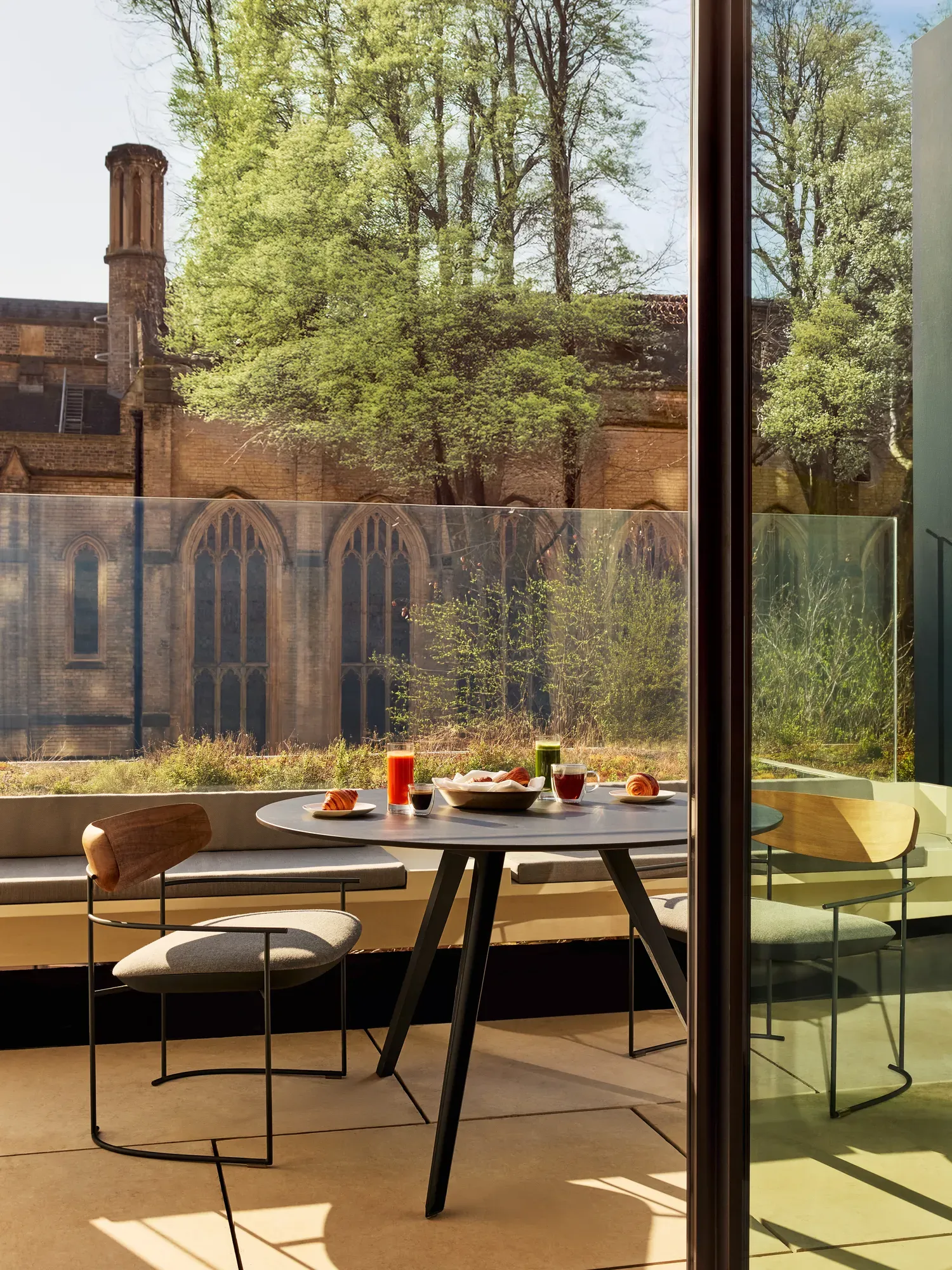 Outdoor table with croissants, juice, and coffee overlooking historic stone church and greenery.