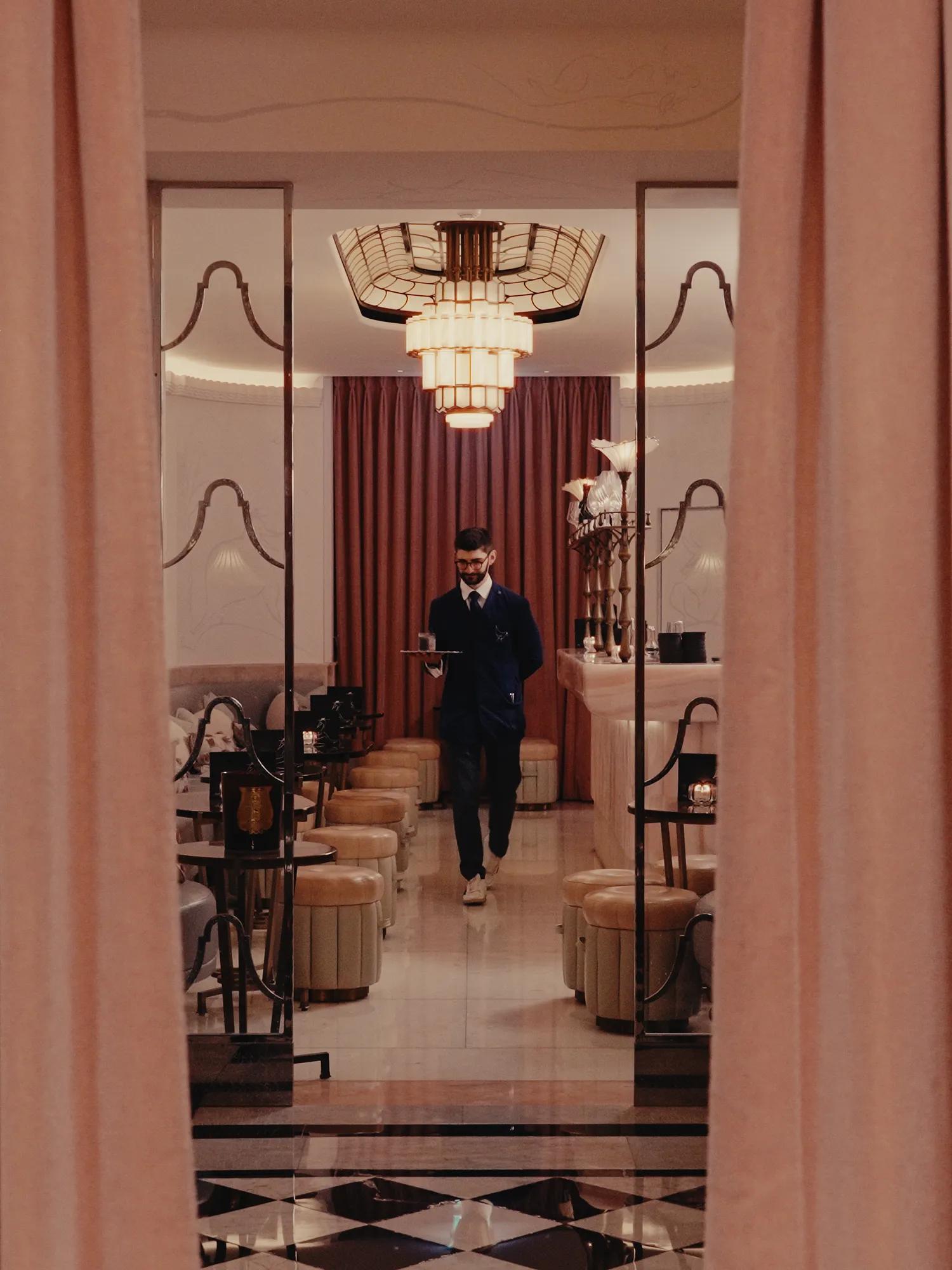 A server carrying a tray walks through an elegant Art Deco-inspired dining room, framed by soft lighting, polished marble floors and warm blush-toned interiors.