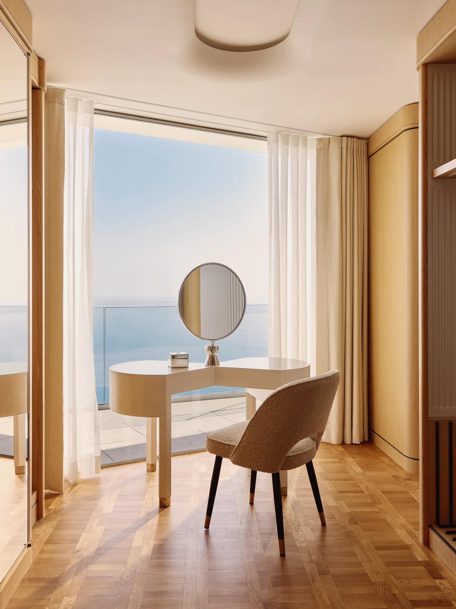 Coiffeuse élégante avec miroir rond et chaise près de baies vitrées, donnant sur une terrasse avec vaste vue sur la mer.