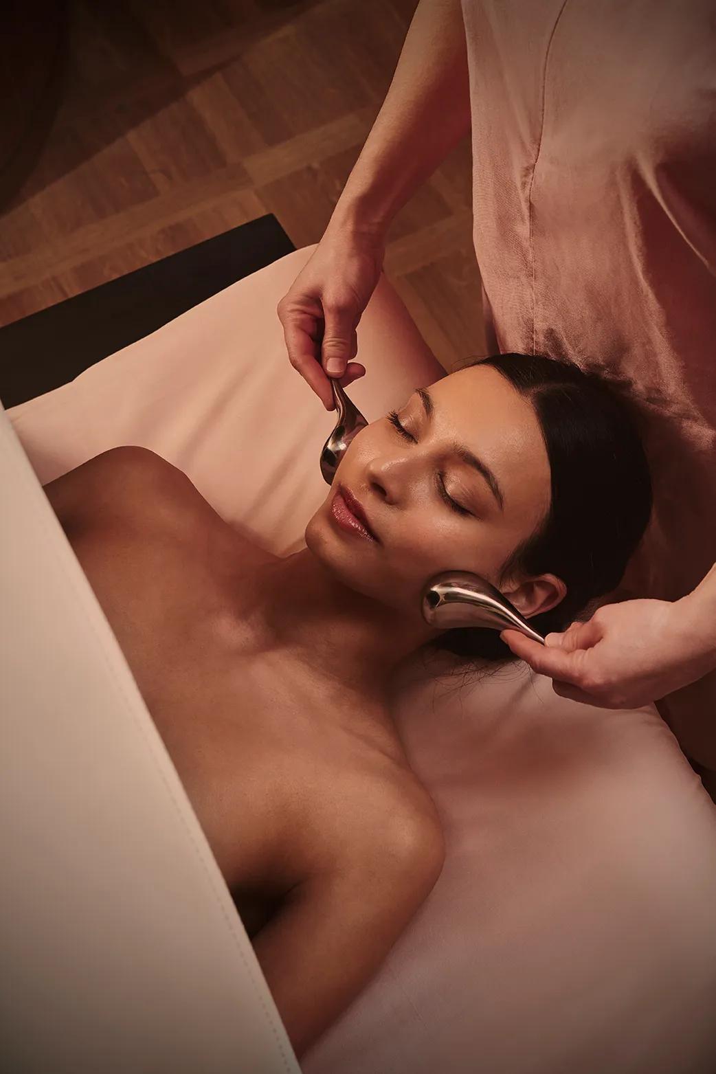 Spa therapist performing a facial massage with cooling metal rollers on a guest’s cheeks