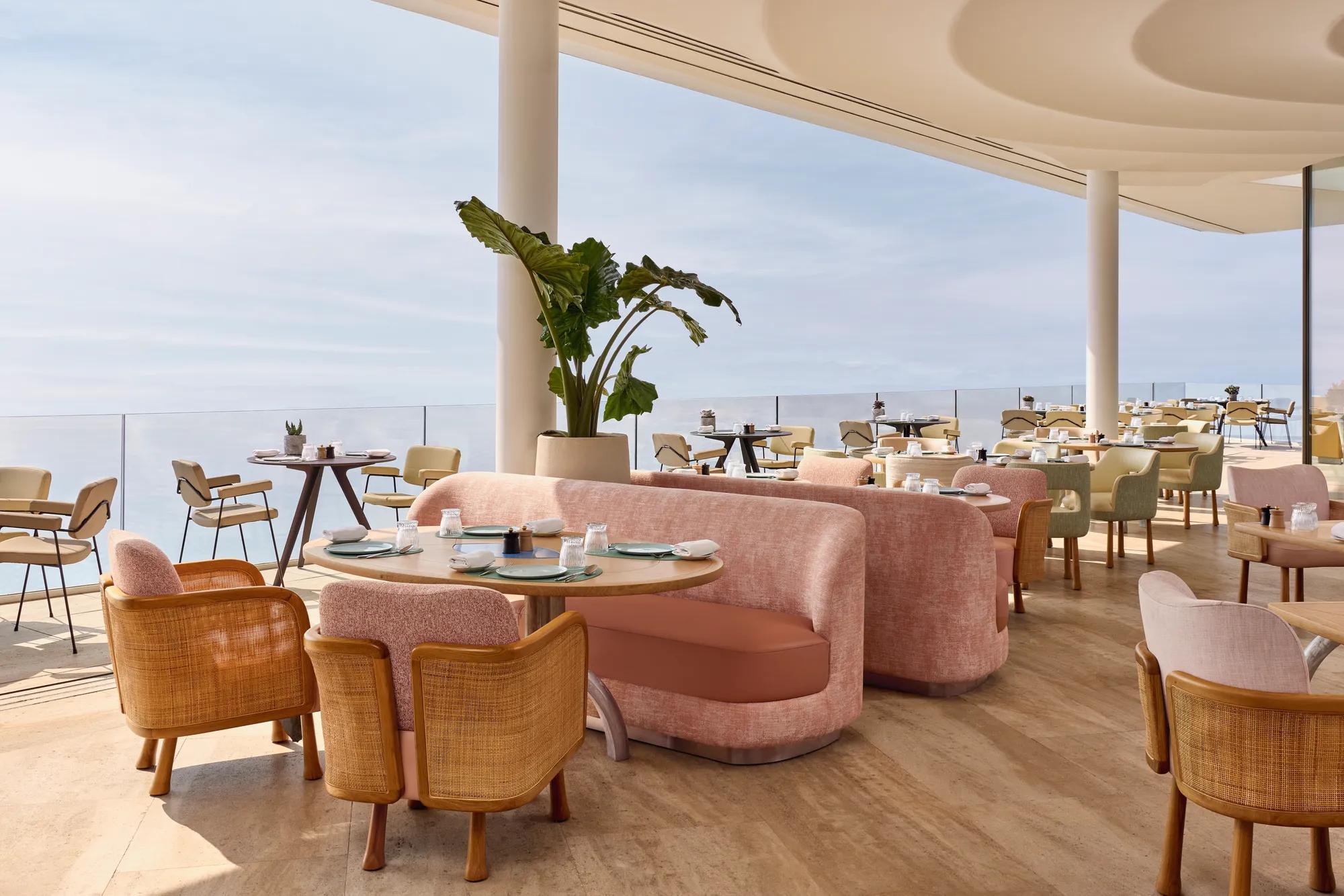 Elegant restaurant interior with pink sofas, wooden chairs, and sea views through floor-to-ceiling windows, featuring soft natural light and modern décor.