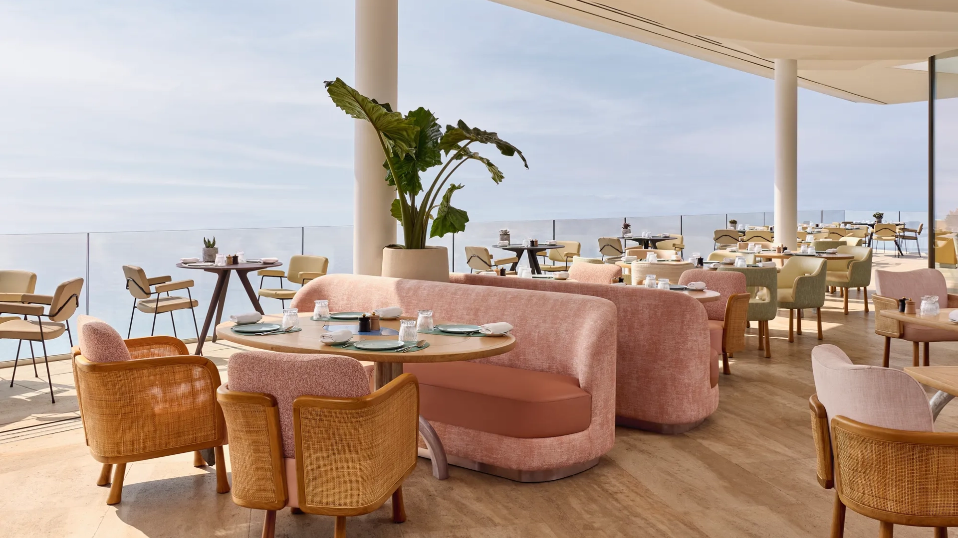 Intérieur élégant de restaurant avec canapés roses, chaises en bois et vue sur la mer à travers de larges baies vitrées, baigné de lumière naturelle.