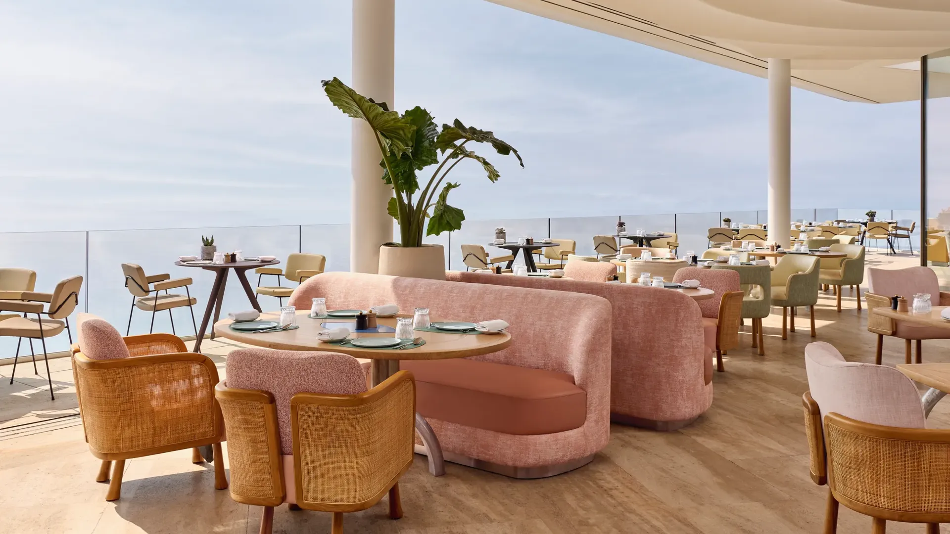 Intérieur élégant de restaurant avec canapés roses, chaises en bois et vue sur la mer à travers de larges baies vitrées, baigné de lumière naturelle.