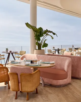 Intérieur élégant de restaurant avec canapés roses, chaises en bois et vue sur la mer à travers de larges baies vitrées, baigné de lumière naturelle.