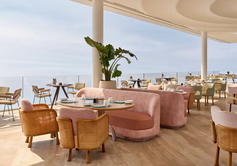 Intérieur élégant de restaurant avec canapés roses, chaises en bois et vue sur la mer à travers de larges baies vitrées, baigné de lumière naturelle.