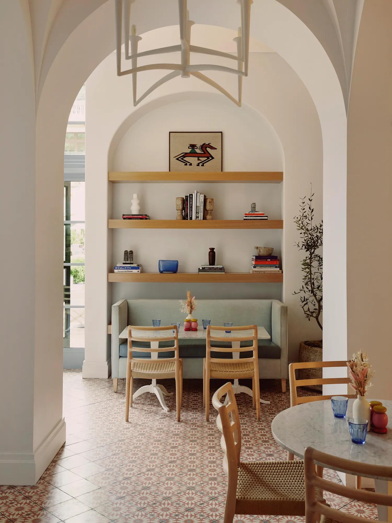 Café alcove with booth, two chairs, and shelves holding books, art, and vases beneath a vaulted arch.