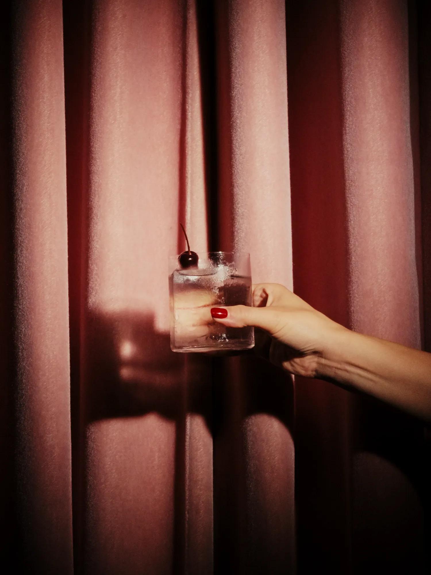 A hand holding a cherry-garnished cocktail against rich velvet curtains, illuminated by warm, atmospheric lighting.